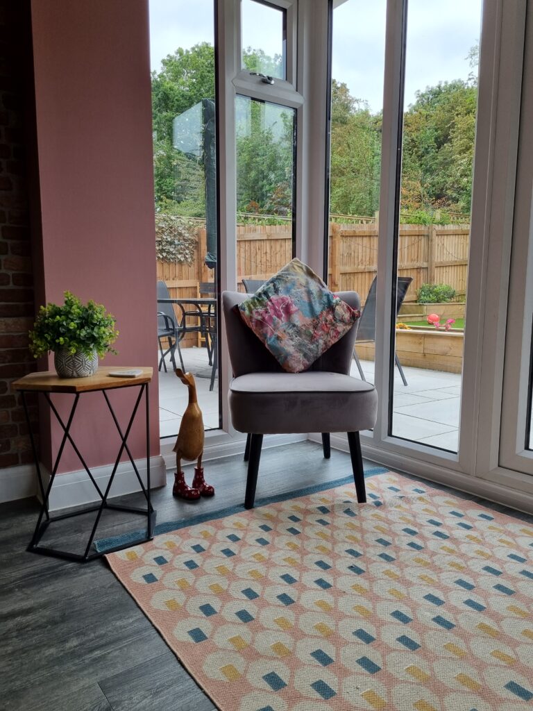 A cushioned chair with a floral pillow sits beside large windows overlooking a patio and garden. A small side table with a potted plant and a standing duck decoration are nearby.