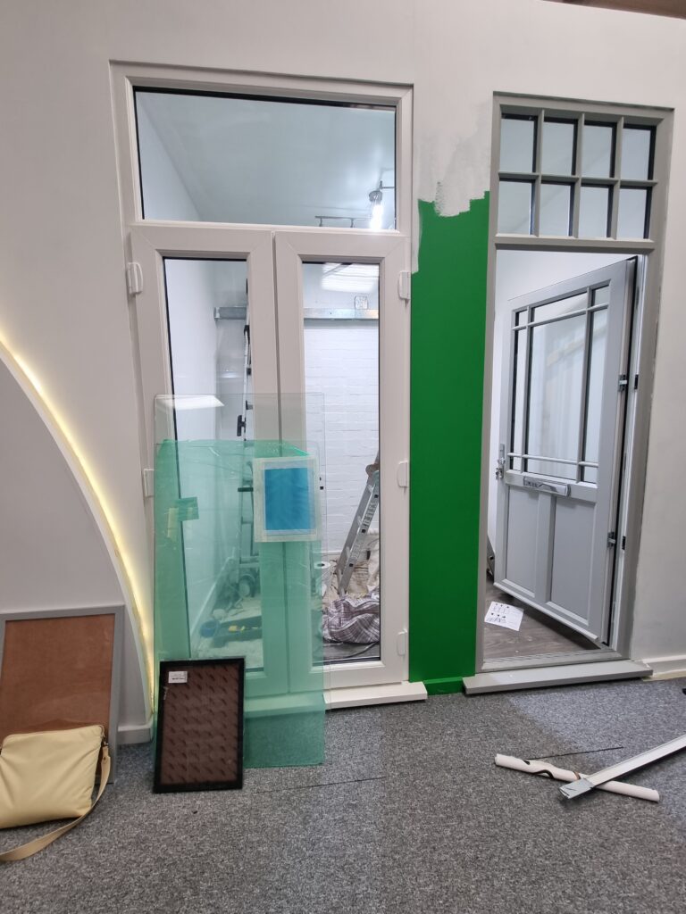 A white-framed window and green-painted wall partially obscure an interior room, with construction tools and materials, including glass panes and a folded ladder, scattered on a carpeted floor.