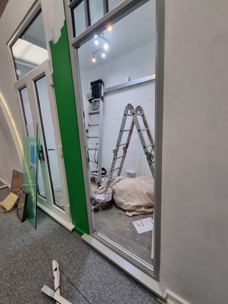 A partially painted room with a green and white wall; inside, a folded ladder and construction materials are on the floor under bright ceiling lights. Adjacent unfinished area with glass panes and carpet.