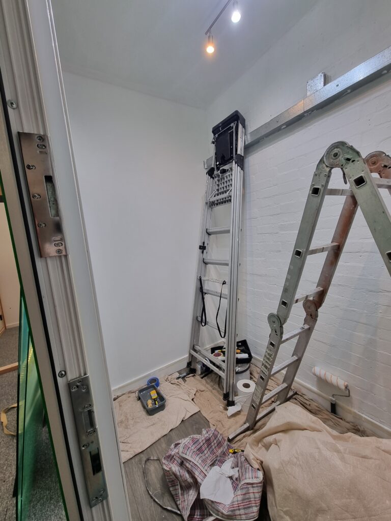 Ladders leaning against a white wall, paint roller on the ground, surrounded by drop cloths and tools, in a small, well-lit room.