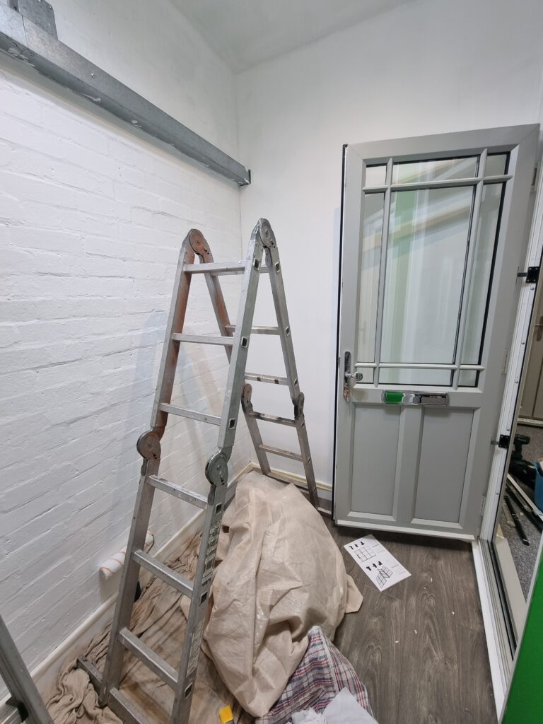 A folded metal ladder leans against a white brick wall, surrounded by drop cloths and tools. An adjacent open door leads to another room with visible laminate flooring.