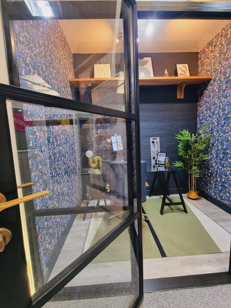 Office interior viewed through open black-framed glass door. A chair and desk are situated on a green and white rug within a small, floral wallpapered room. Shelf holds framed art.