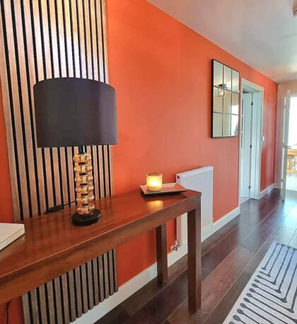 A wooden table with a black lamp and lit candle sits against an orange wall with vertical wood slats, a mirror, and adjoining hallways.