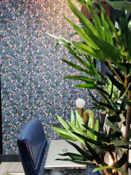 A closed silver laptop and a white lamp sit on a glass desk beside a blue chair, in front of a floral-patterned wall, partially obscured by green plant leaves.