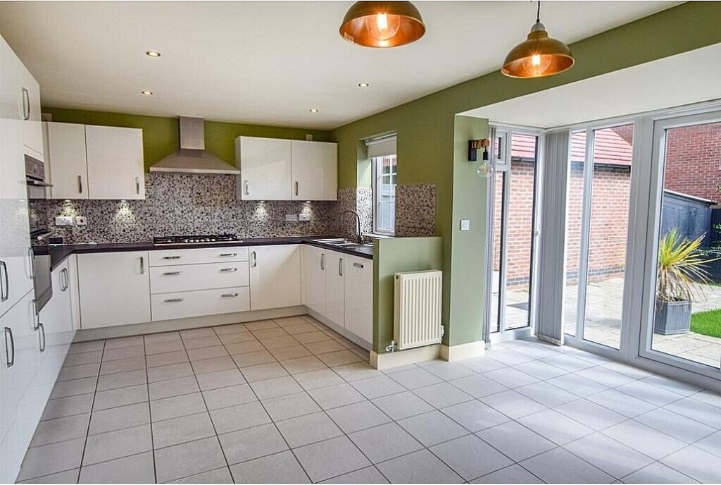 A modern kitchen, featuring white cabinets and stainless steel appliances, is illuminated by overhead lights. Large windows on the right let in natural light, with an outside view of a brick wall and patio.