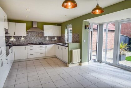 A modern kitchen, featuring white cabinets and stainless steel appliances, is illuminated by overhead lights. Large windows on the right let in natural light, with an outside view of a brick wall and patio.