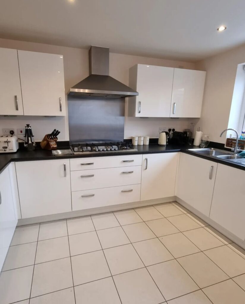 Modern kitchen with white cabinets, a stainless-steel range hood, gas stove, and black countertops. Appliances like a kettle and toaster are on the counter. White tiled floor and neutral walls.