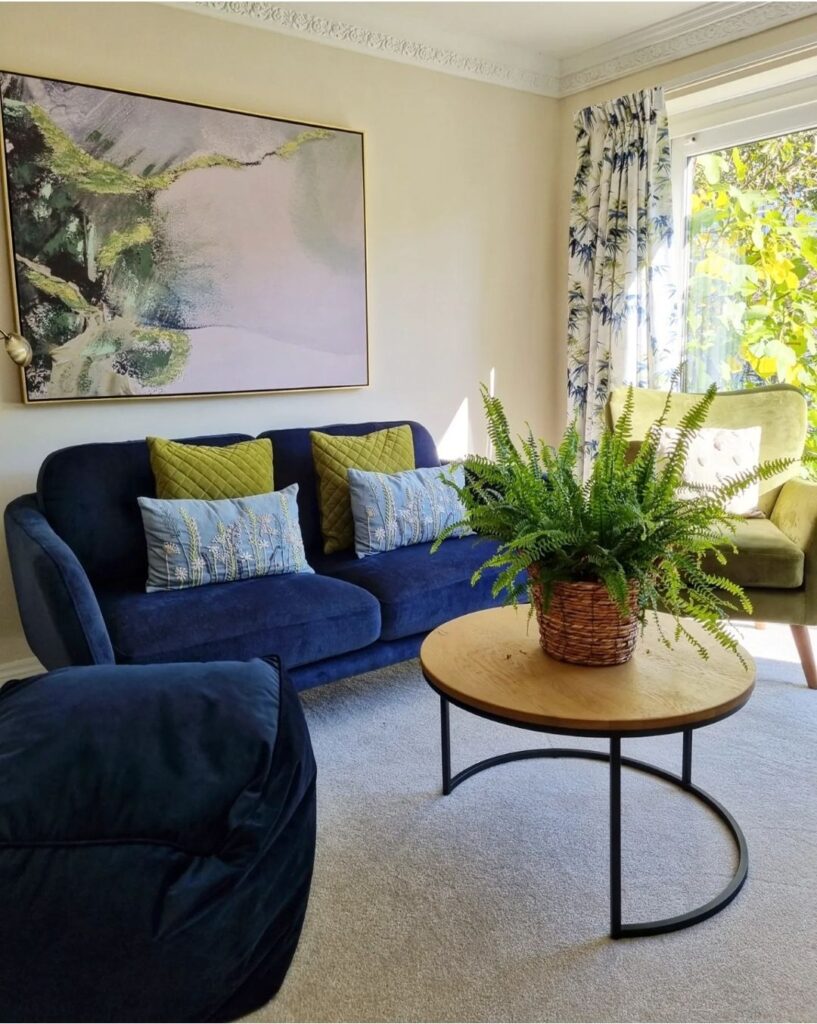 A blue sofa with green and blue cushions, placed against a cream wall with a large abstract painting. A round wooden table with a potted fern sits on a beige carpet.