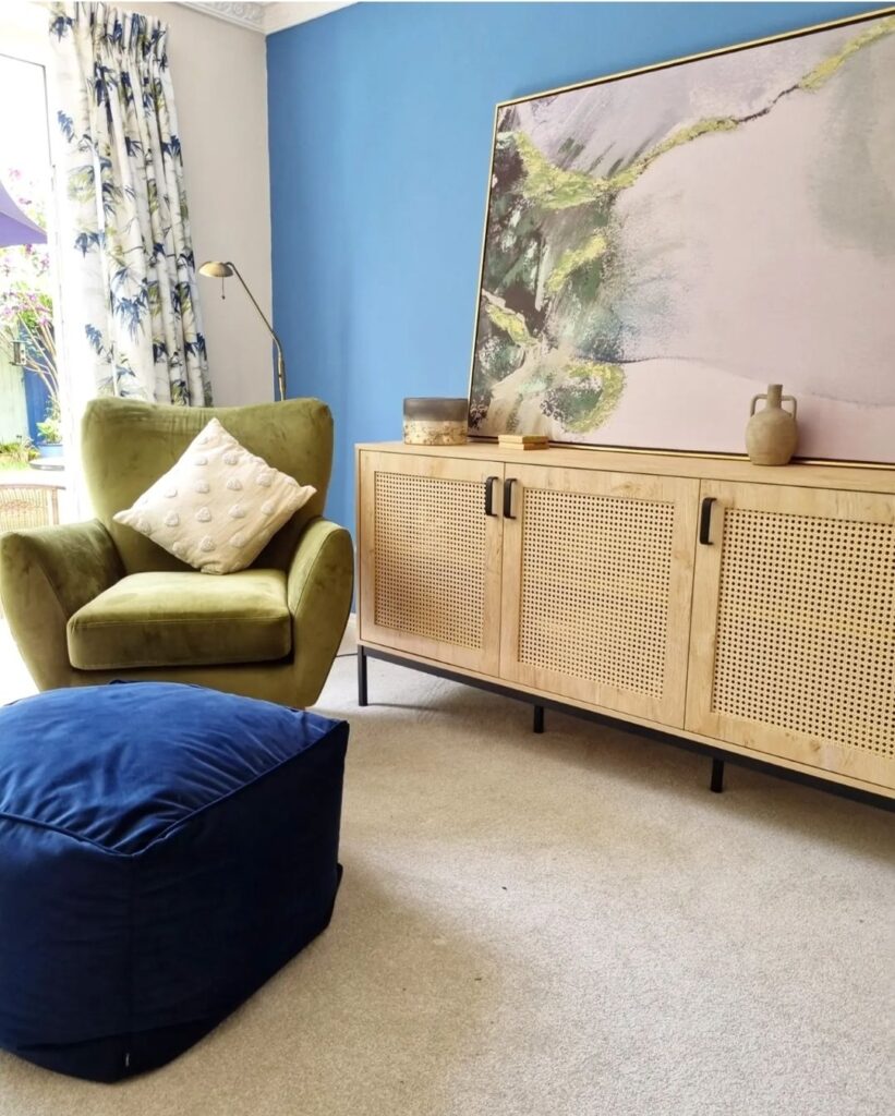Green armchair with white cushion and blue ottoman in a carpeted room beside a wooden cabinet with a large abstract painting leaning against a blue wall.