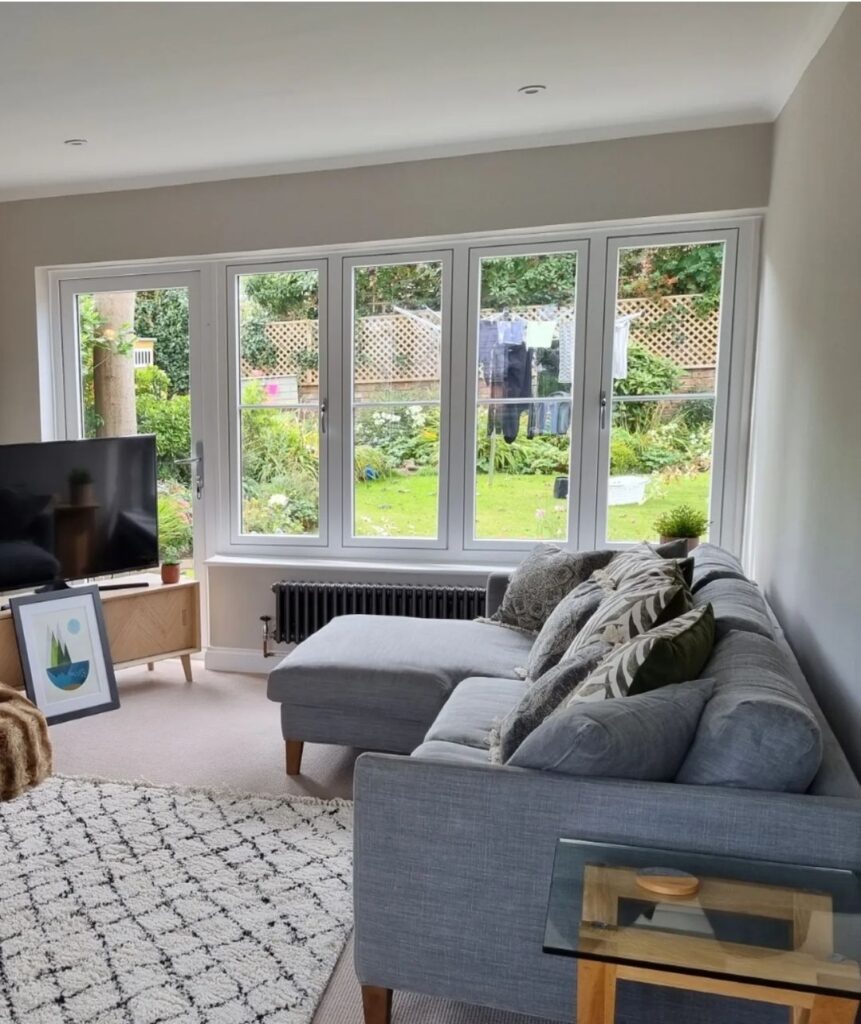 A gray sectional sofa sits in a bright living room, facing a TV. Large windows showcase a lush garden with a wooden fence. A patterned rug and small side table accent the space.