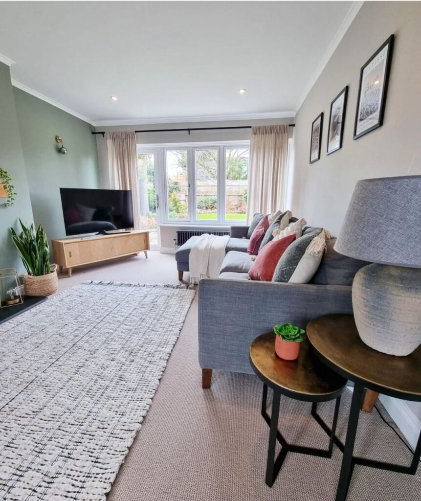 A cozy living room features a gray sofa with colorful cushions, a TV on a wooden stand, a large rug, and windows with beige curtains provide a view of a garden.