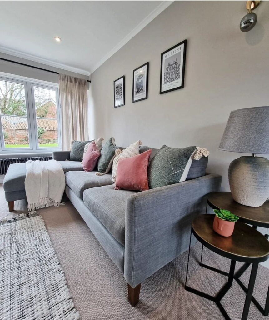 A gray sofa adorned with colorful cushions sits in a well-lit living room beside a window. Three black-framed artworks captioned "Matisse" hang on the light-gray wall above.