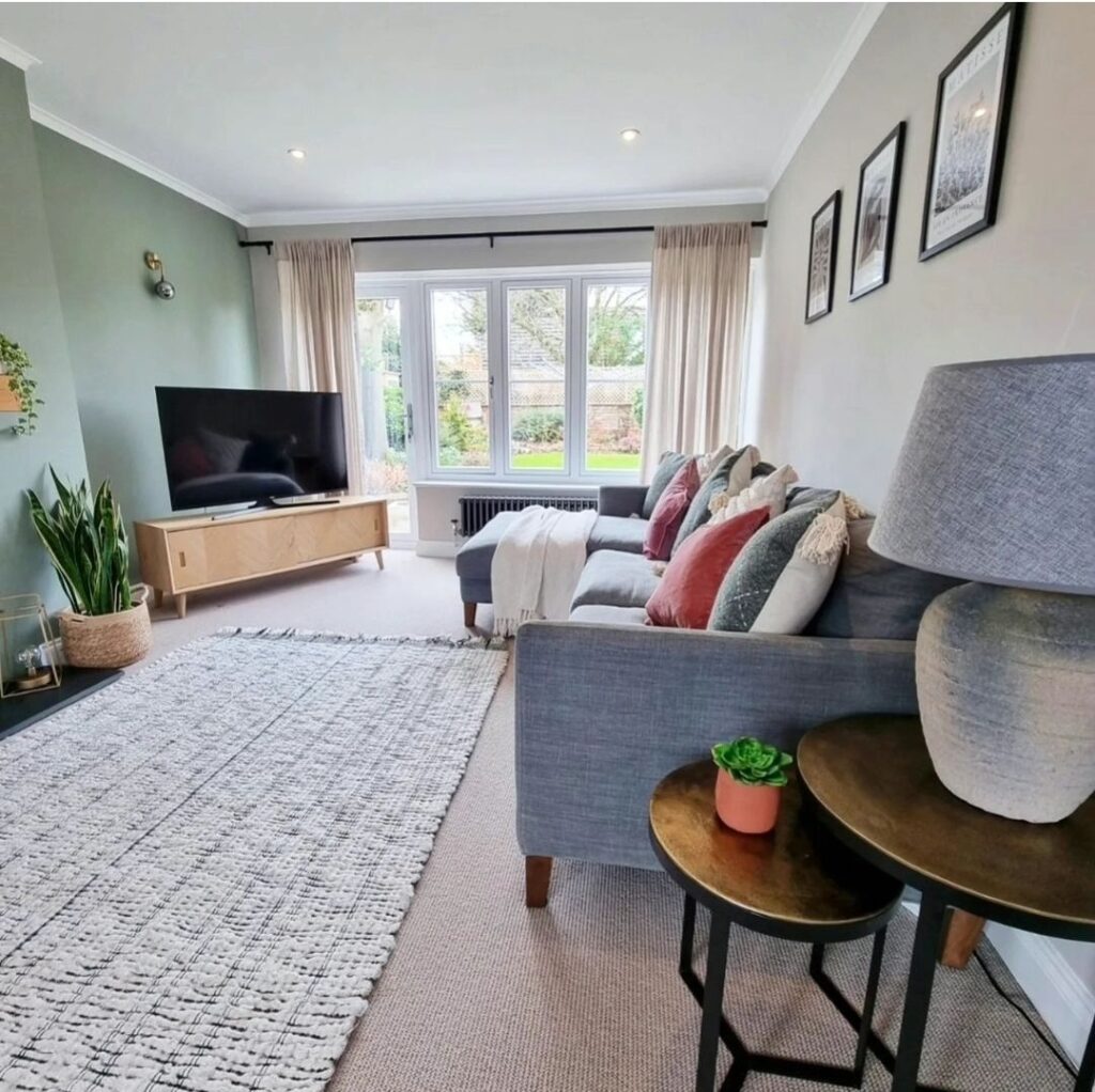 A grey sectional sofa with colorful pillows faces a flat-screen TV on a wooden stand in a well-lit living room featuring a large window, framed pictures, and a grey-patterned rug.