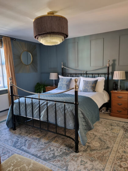 A metal-framed bed with blue bedding and patterned pillows sits in a well-lit bedroom with teal walls, wooden nightstands, a decorative ceiling light, and a sunburst mirror above a rug.
