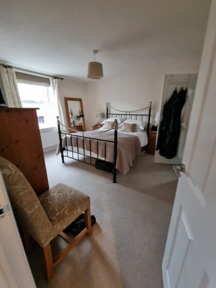 Iron bed with pillows and a beige throw in a carpeted bedroom, surrounded by a dresser, large mirror, armchair, and window with curtains. Clothing hangs in an open closet door.