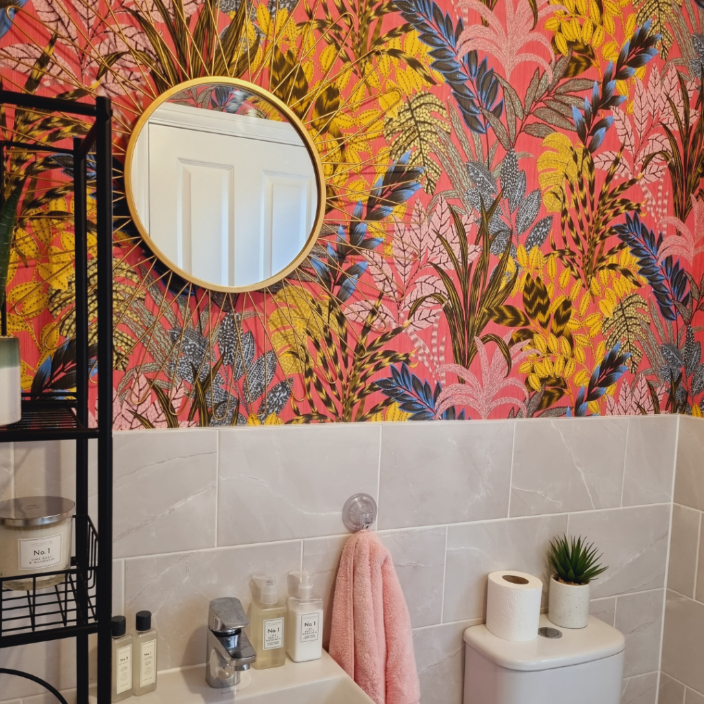 Circular mirror with sunburst gold frame reflecting a white door, mounted on a vibrant botanical-patterned pink wall; adjacent to a marble-tiled sink area with toiletries and a small plant.
