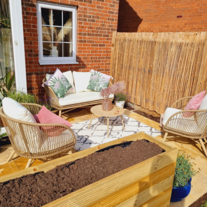 A cozy patio setup features wicker chairs and a sofa with decorative cushions surrounding a small round table, set against a brick wall and a bamboo privacy screen.