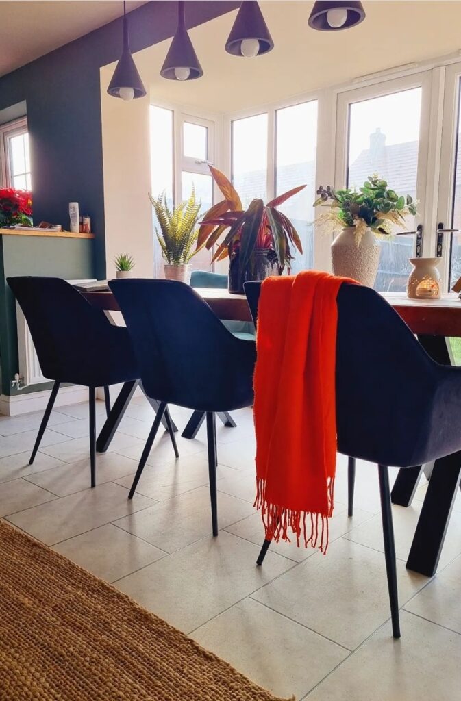 Four black chairs surrounding a wooden dining table adorned with plants, a vibrant orange throw draped over one chair, in a brightly lit room with large windows and hanging lights.