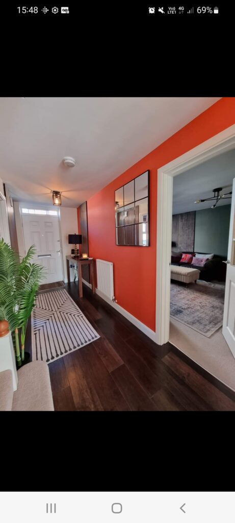 A hallway features a bright reddish-orange wall with a grid-style mirror, a small console table with a black lamp, and a white front door. Adjacent room with cozy living area visible.