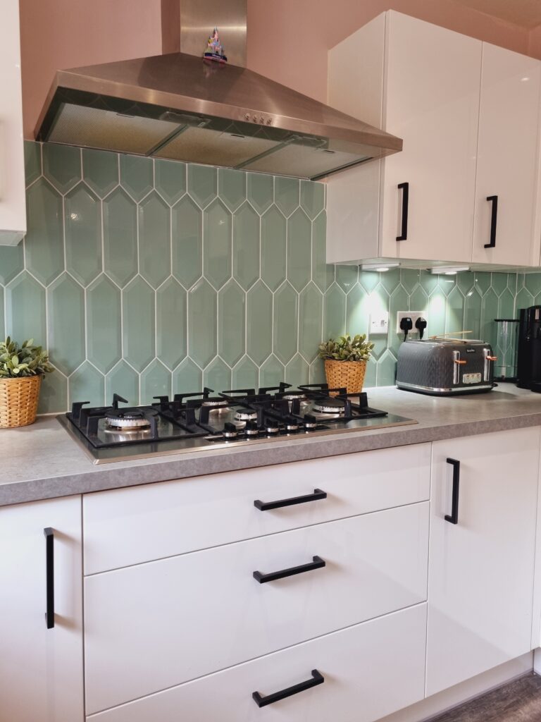 Stainless steel gas stove on a grey countertop with white cabinets and green hexagonal-tiled backsplash in a modern kitchen; potted plants and toaster are on the countertop.