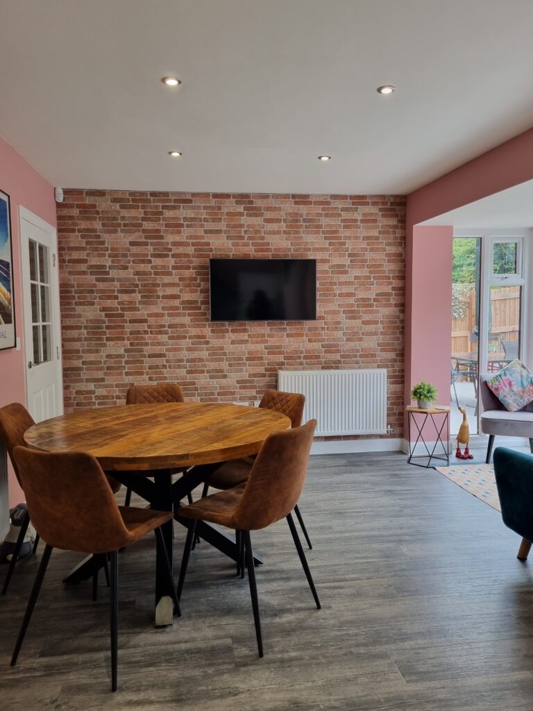 A wooden round dining table with four brown chairs sits in a modern room with a brick accent wall, a wall-mounted TV, pink walls, windows, and contemporary decor.