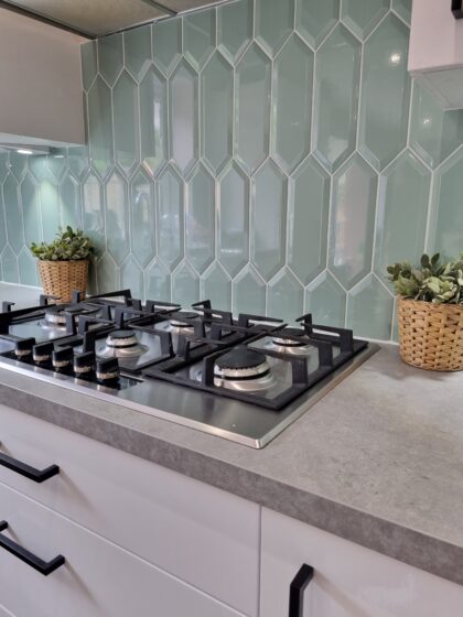 A modern gas stove is set against a green, hexagonal-tiled backsplash with two potted plants in woven baskets flanking it on a gray kitchen countertop.