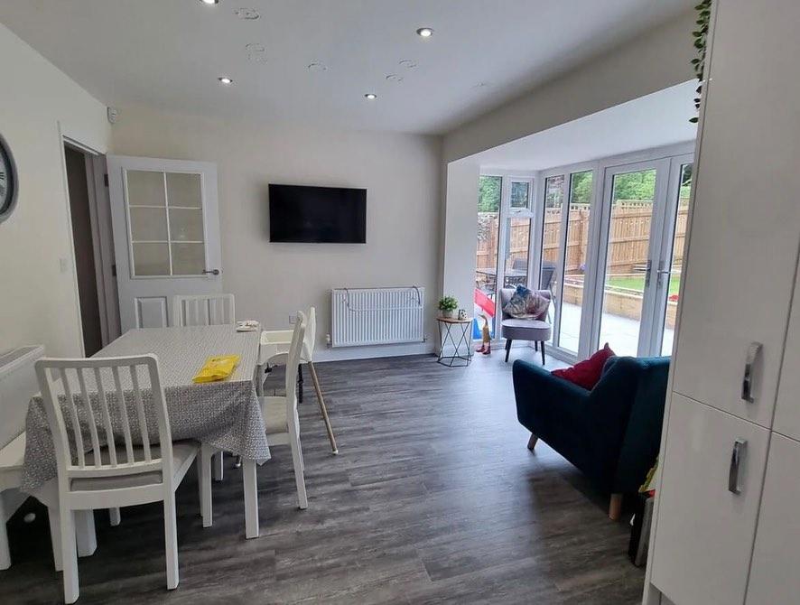 A modern dining room with a table and chairs, situated beside a living area with an armchair, adjacent to glass doors leading to a patio. Walls are beige with a mounted TV.