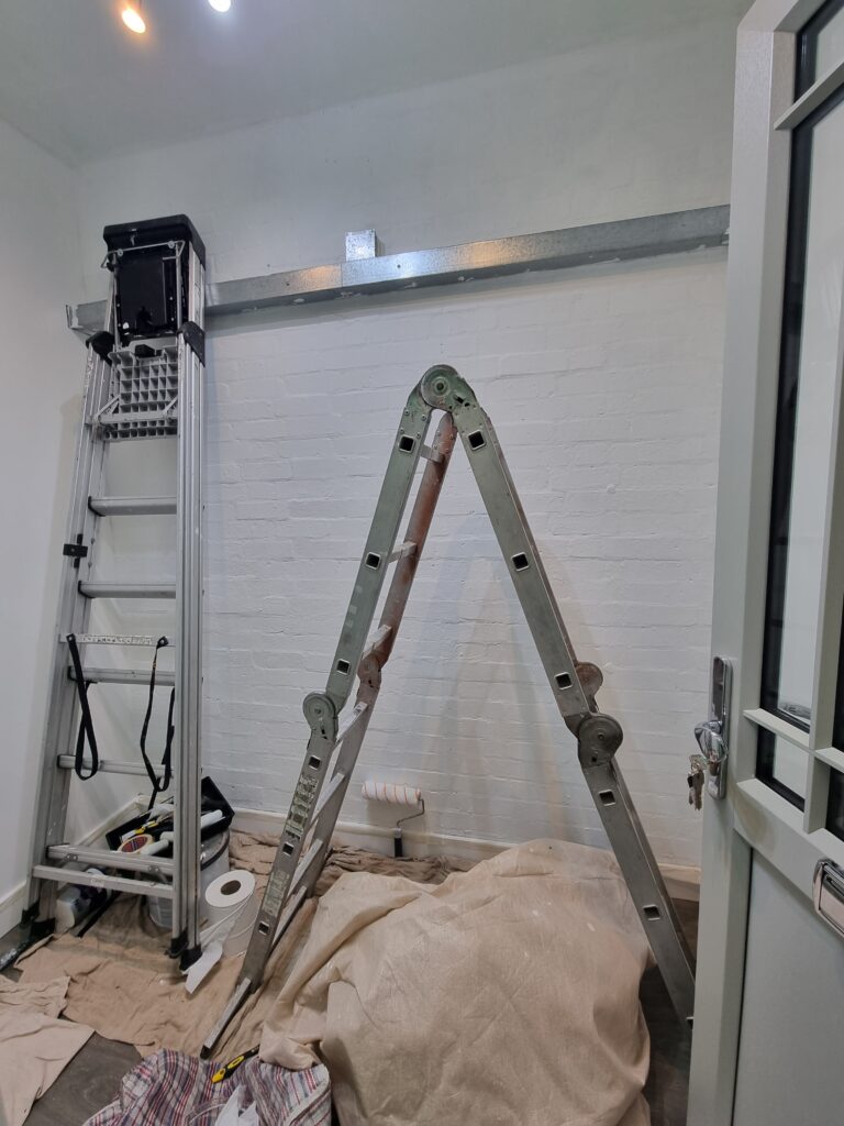A metal ladder stands open alongside a taller folded step ladder against a white brick wall. Various tools and materials, including a roller and cloths, are scattered on the floor.