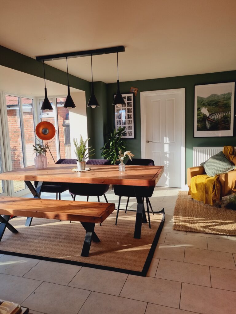 Wooden dining table with matching bench and purple chairs sits on a beige rug, near potted plants, floor lamp, and mustard-yellow draped armchair, under black pendant lights in a sunlit room.