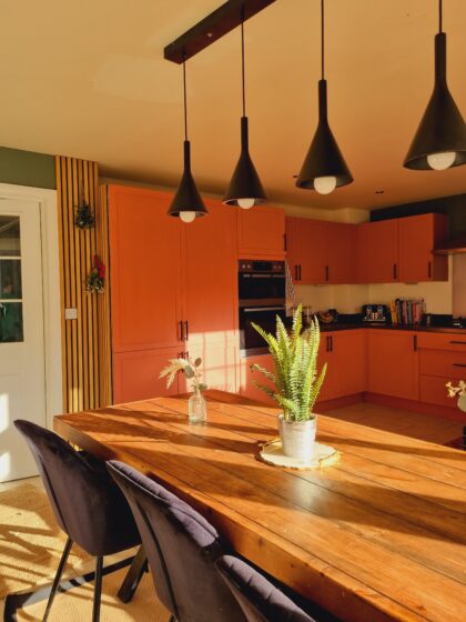 A wooden dining table with a potted plant and vase sits under black pendant lights, in a warm kitchen with orange cabinets and adjacent white door, basking in sunlight.