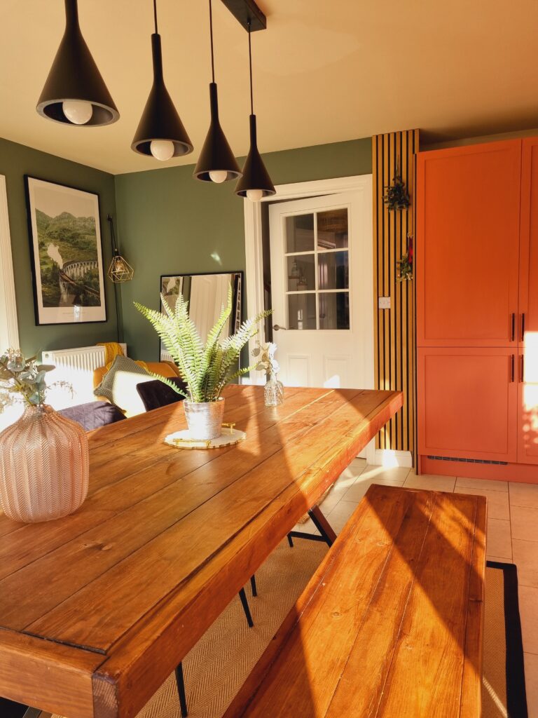 A wooden dining table with a potted fern centerpiece is flanked by matching benches under black pendant lights in a sunlit room with green walls, framed art, and an orange cabinet.