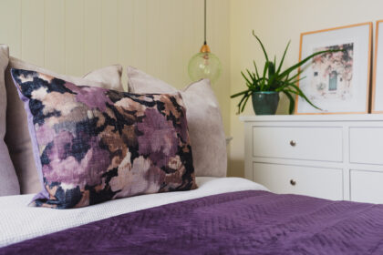A floral-patterned pillow rests on a bed with purple bedding. Nearby, a white dresser holds a potted plant and framed art. A green hanging lamp is adjacent.