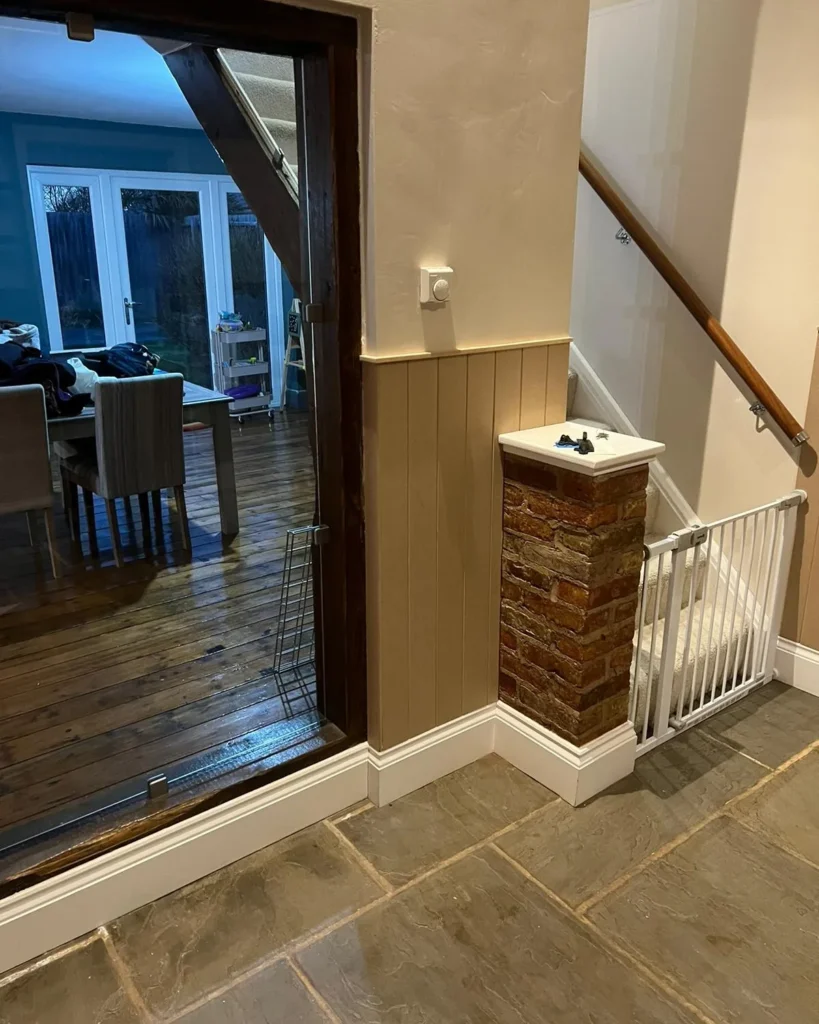 A stairway with a white baby gate is adjacent to a wooden-framed glass door leading to a dining area with wooden floors and white chairs around a table.