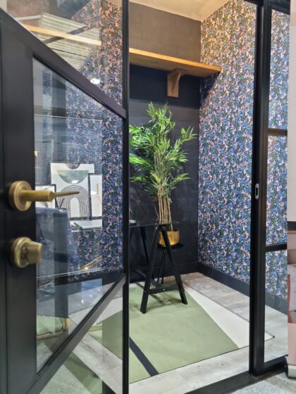 A potted plant sits on a black stand in a small room with floral wallpaper, a green rug, and a wooden shelf. The room is visible through a glass door with a brass handle.