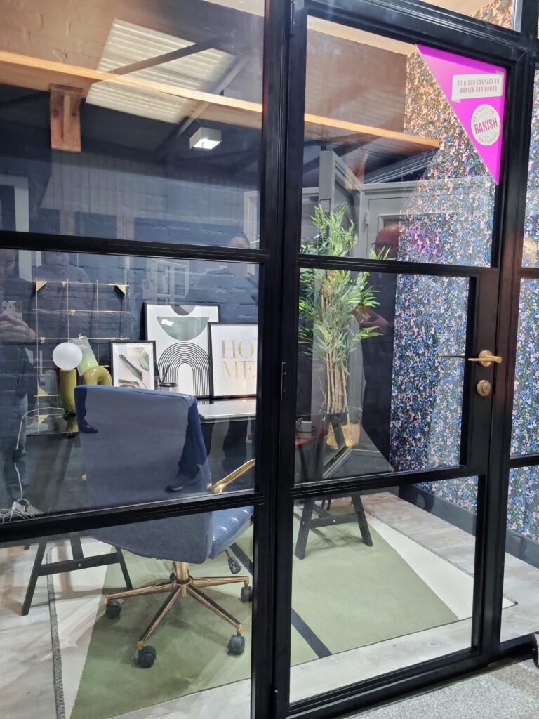 A modern office setting features a blue swivel chair on a green rug, with framed artwork and potted plants against a black wall. Text on a pink triangle reads “JOIN OUR CRUSADE TO BANISH BAD ODORS.”