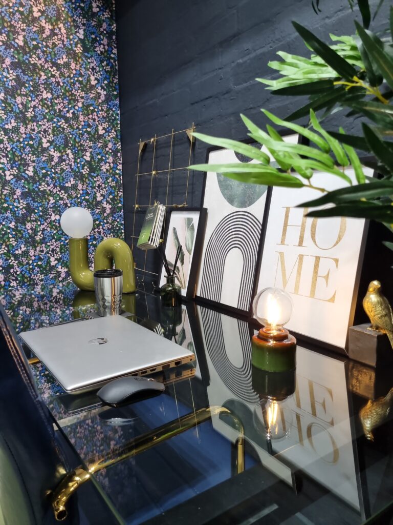 Laptop and mouse on a glass desk, surrounded by decorative items including lamps, plants, and framed artwork in a room with a floral-patterned wall and a dark-painted brick wall.