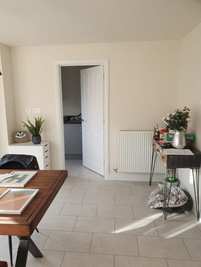 Wooden table with framed art sits in a tiled room; a plant and files are on an adjacent cabinet, and an open door leads to another room.