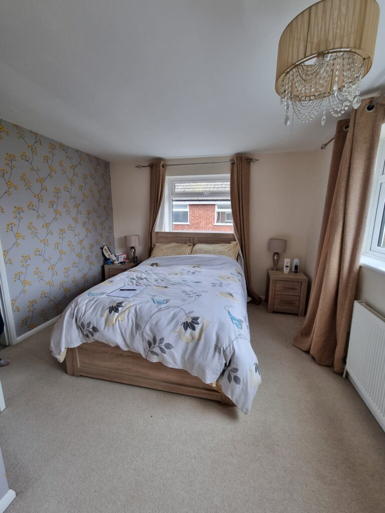 A bed adorned with a floral duvet sits against a floral wallpapered wall in a cozy, beige-themed bedroom with large windows and decorative light fixtures.