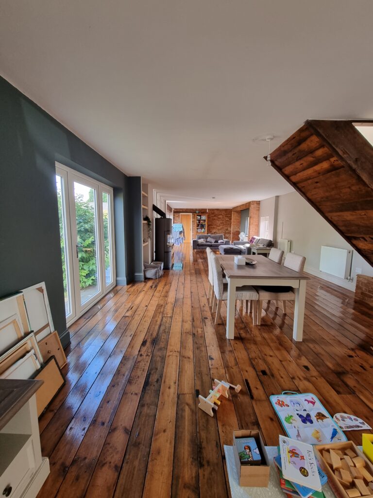 Wooden dining table with cream chairs and scattered toys, located in a long room with large windows and wooden flooring, leads to a cozy seating area with sofas and bookshelves.