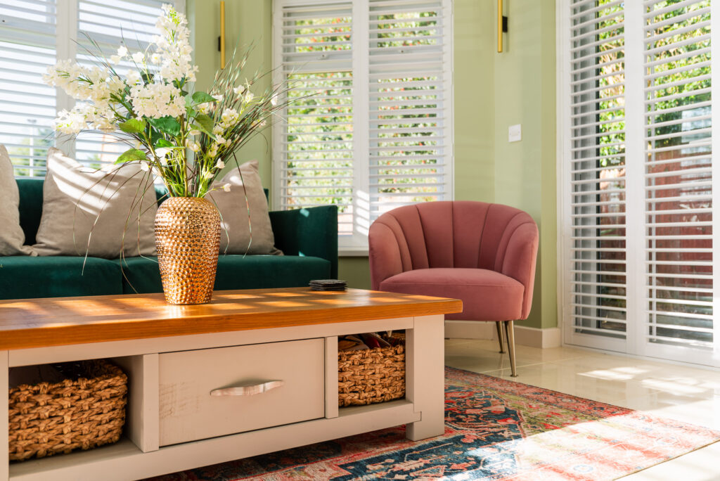 A golden vase with white flowers sits on a wooden coffee table in a sunlit living room with a green sofa, pink armchair, colorful rug, and white plantation shutters.