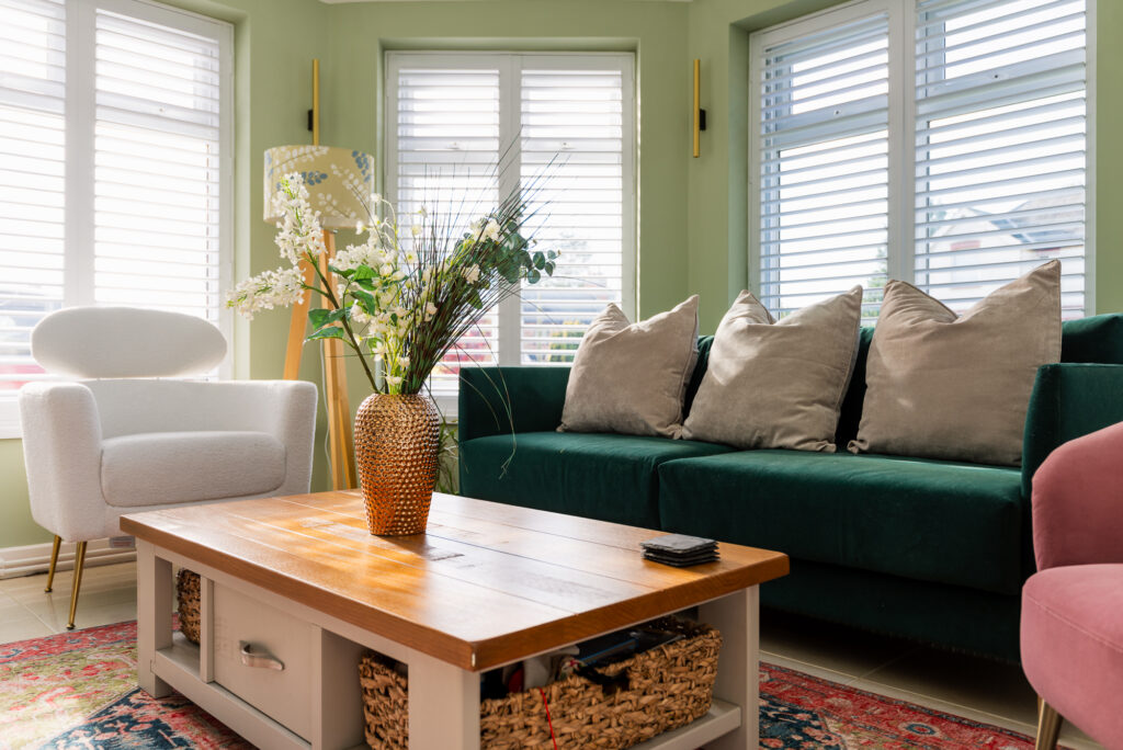 Vase of flowers on a wooden coffee table, surrounded by a white armchair, green sofa with cushions, and windows with blinds in a brightly lit living room.