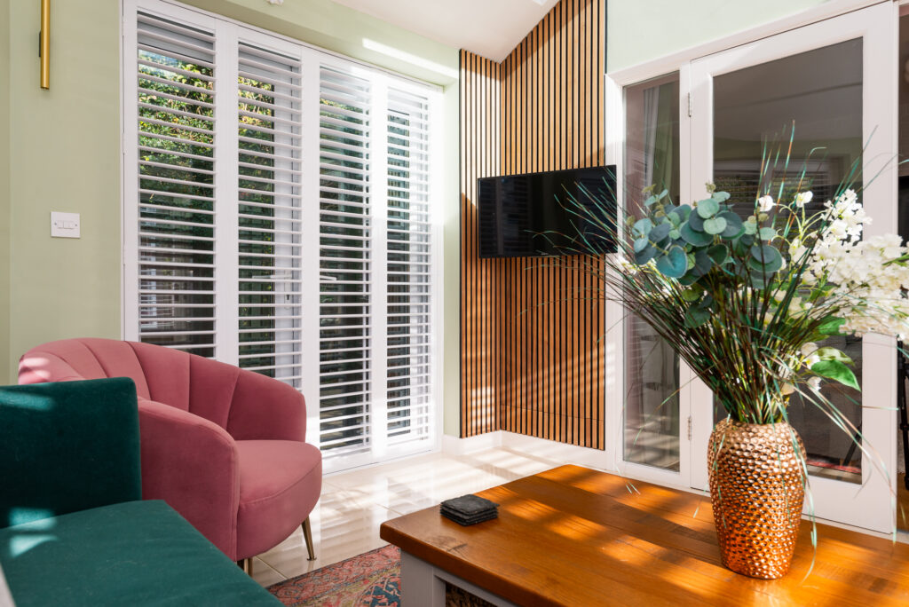 Pink armchair and teal sofa sit in a sunlit room with white plantation shutters, wooden paneling, and a mounted TV. A wooden coffee table holds a gold vase filled with greenery and white flowers.