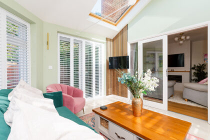 A comfortable living area with a green sofa, pink chair, wooden coffee table with flowers, and large windows with white shutters; adjacent room visible through glass doors.