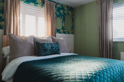 A neatly made bed with gray and teal pillows and a quilted teal blanket is positioned against large windows with shutters and beige curtains in a room with green and floral wallpaper.