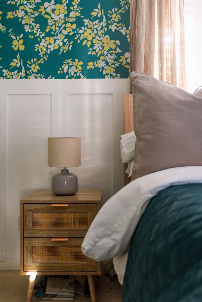 A wooden bedside table supports a gray lamp and sits next to a bed with gray and white pillows, under a green floral wallpaper and beside beige curtains.