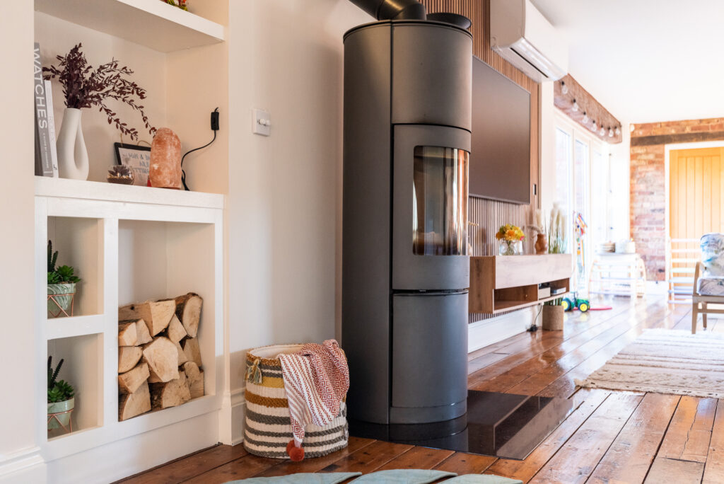 A black cylindrical wood-burning stove stands in a modern, cozy living room with wooden flooring, next to a built-in shelf with firewood, books, and plants.