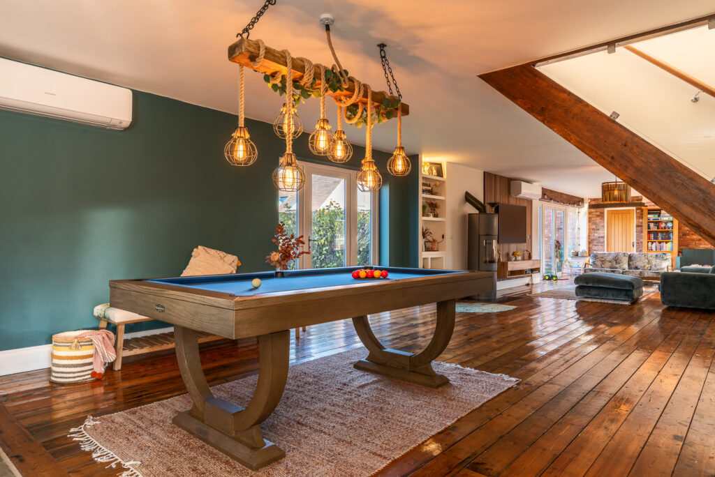A wooden pool table with blue felt is under rustic pendant lights in a spacious, stylish living room featuring a green accent wall, wooden flooring, and a seating area with modern furniture.