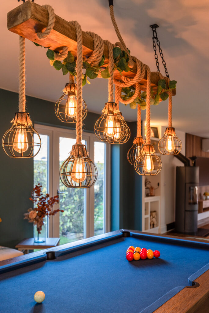 A pool table with balls is positioned beneath a rustic chandelier made of metal-caged light bulbs hanging from ropes, in a well-lit room with large windows and cozy decor.