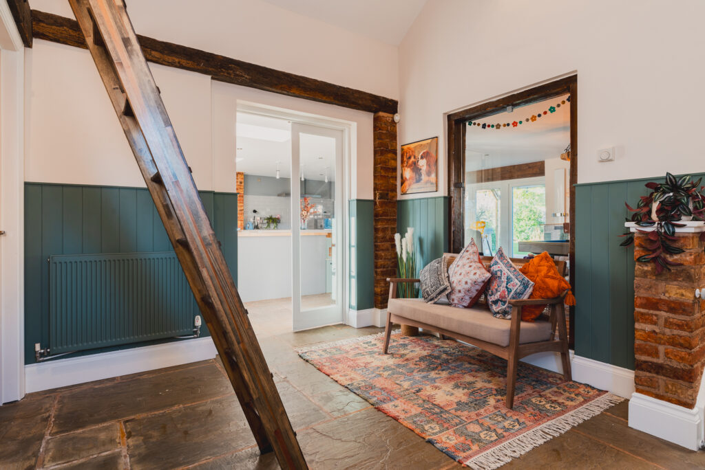 Wooden ladder leaning against a wall with green paneling, colorful rug, cushion-filled bench, floral artwork, potted plant, and adjacent open doorways leading to bright kitchen and living spaces.