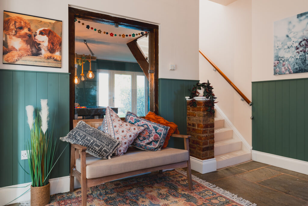 Bench covered with assorted pillows, placed against a green-paneled wall beneath a dog portrait, next to staircase with framed floral art and plant decor; adjacent to a room with hanging lights.